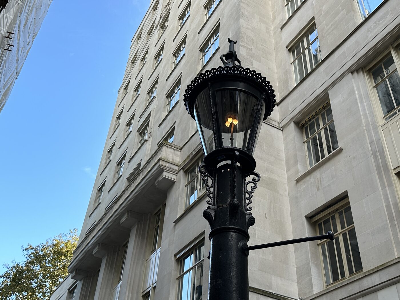 An ornate cast iron gas lamp burning in bright daylight on Carting Lane, London, with a pale stone building behind it